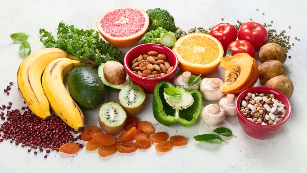 A spread of healthy foods high in potassium, including bananas, avocado, potatoes, dried apricots, beans, oranges, and assorted vegetables