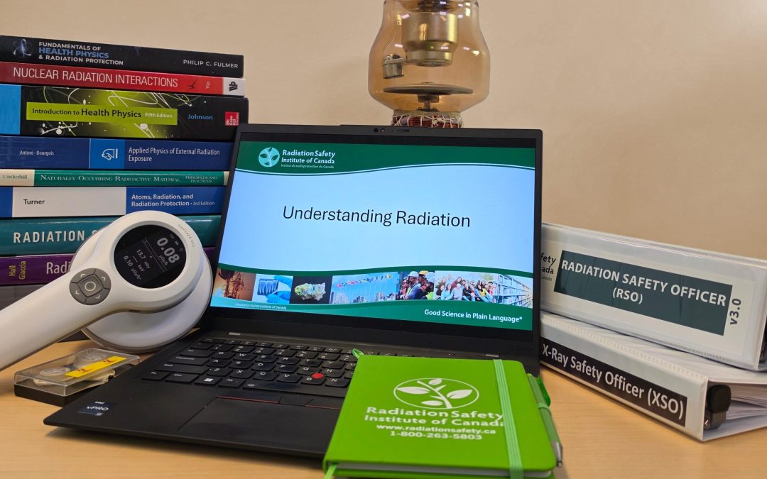 Laptop with "Understanding Radiation" displayed, stack of radiation protection reference materials behind it to the left, a survey meter in front, a Radiation Safety Institute of Canada notepad on the front right, an x-ray tube behind middle, and an RSO binder on top of an XSO binder behind to the right.